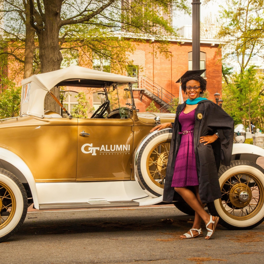 Avery posing in her grad cap and gown in front of a Wreck car with "GT Alumni" written on it