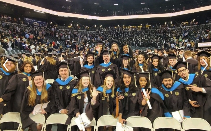 A large group of recent graduates smiling and cheering in their caps and gowns on the floor of the basketball arena